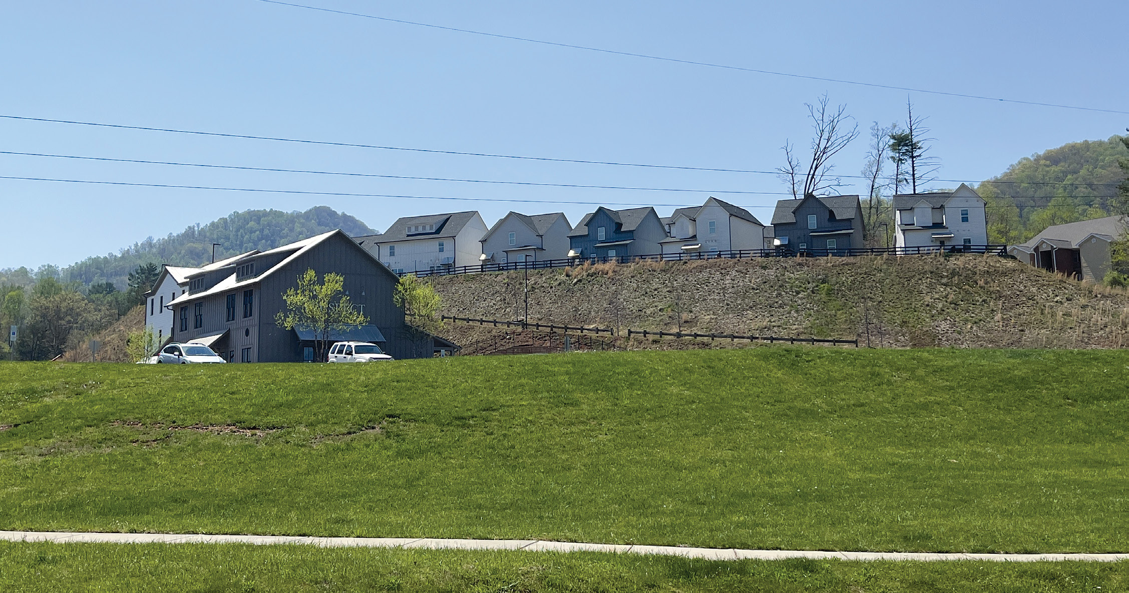 A grassy hillside with a multifamily building in the foreground and a row of houses behind it on higher ground.