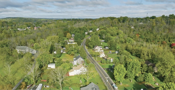  Aerial view of a rural town featuring residential buildings interspersed with trees, green landscape, and a road going down the middle..