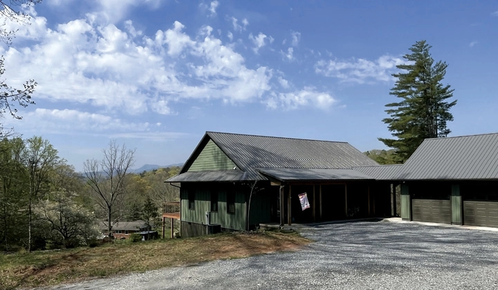 A green one-story home with a gravel driveway overlooking wooded hills.