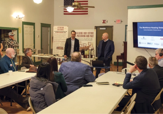 A meeting with participants seated around long tables, with two men presenting in front of a screen titled "The Workforce Housing Solution."