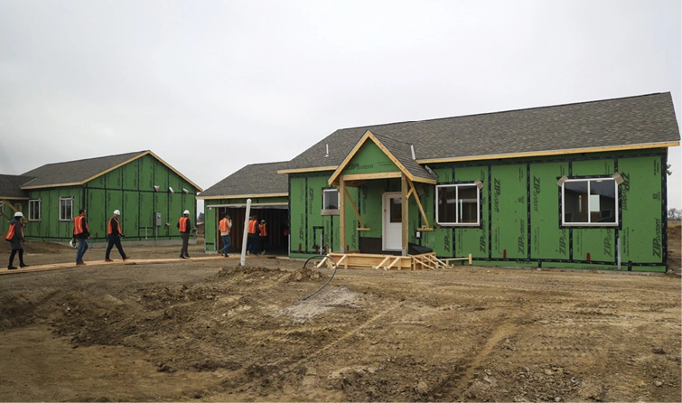 Construction site with partially built houses and a line of workers in orange vests walking into a house.