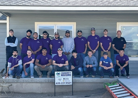 Two rows of people posing in front of a gray, single-story house with roof only partially visible and a "Community Housing" sign in the dirt in front of them.