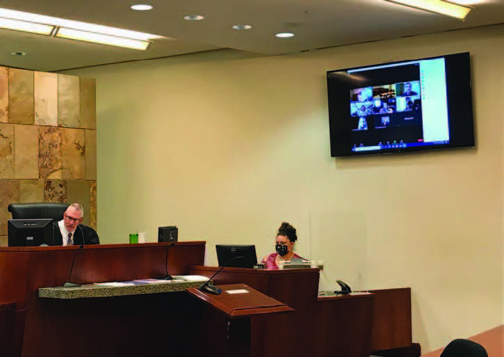View of a courtroom showing the judge and stenographer and people on a television screen mounted on a wall.