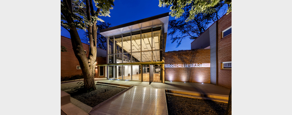 Photograph of the front façade of a modern brick and glass building in the early evening, with a wall sign reading “Dorchester Art.”