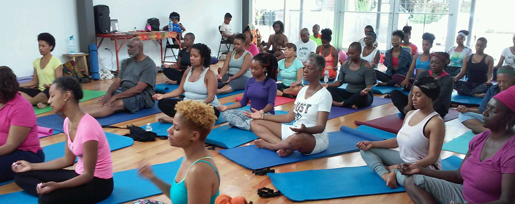 Photograph of approximately 30 people sitting cross-legged on yoga mats in a large, wooden-floored room.