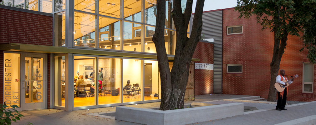 Photograph taken at dusk of the two-story glass front of a modern brick building, with workshop participants inside the building and a guitar player standing outside.