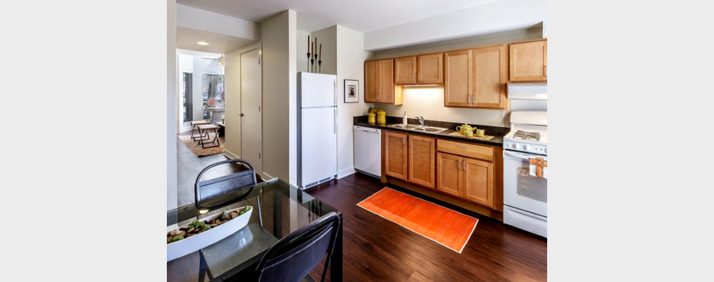 Photograph of the appliances, counter and cabinets, and table in a kitchen, with a wide passageway to the living room in the background.