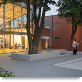 Photograph taken at dusk of the two-story glass front of a modern brick building, with workshop participants inside the building and a guitar player standing outside.