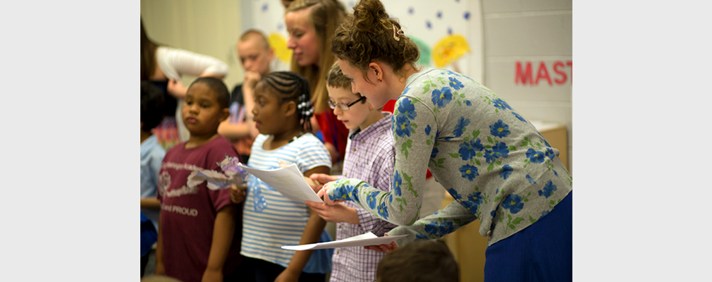 Photograph of two university students helping three children during a rehearsal for a school performance.