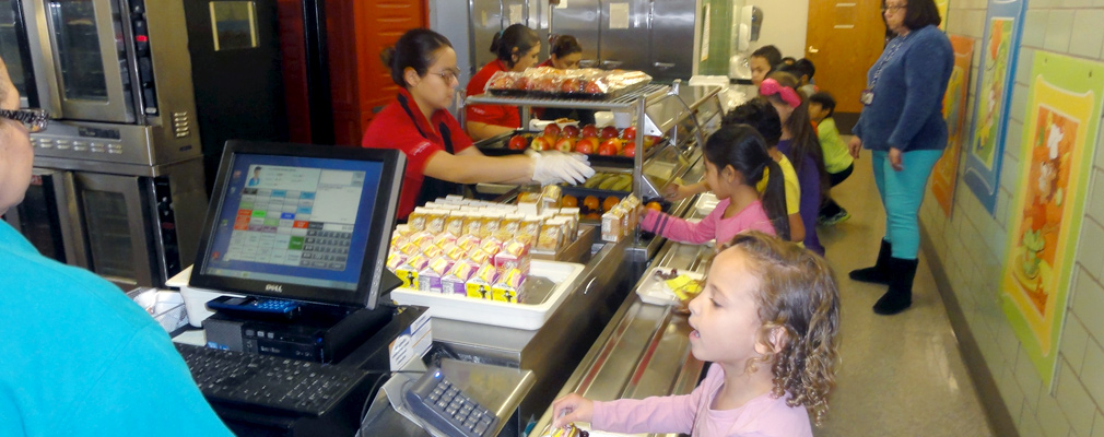 Photograph of eight children receiving an entrée, fresh fruit, and milk at a cafeteria line staffed by four adults.