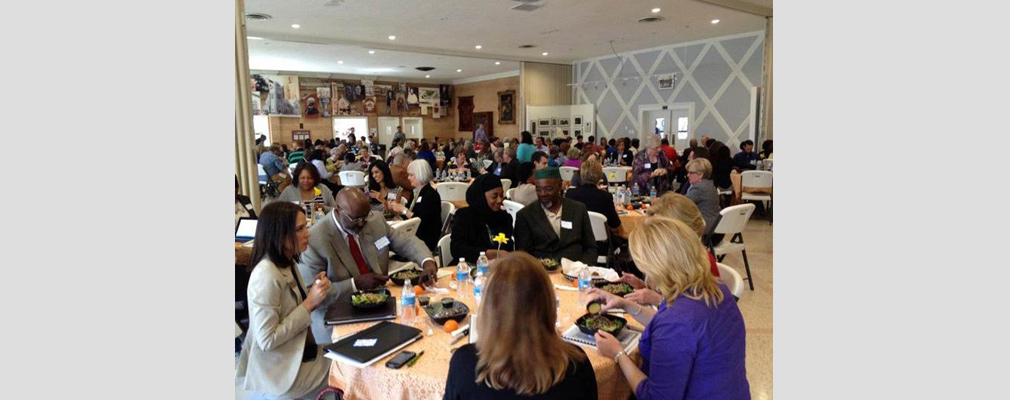 Photograph of dozens of attendees eating lunch at round banquet tables in a large meeting room.