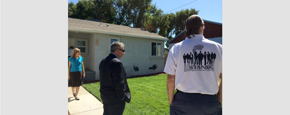 Photograph of three people, one of whom is wearing a shirt with a STAND logo on the back, standing in front of a single-family detached house.