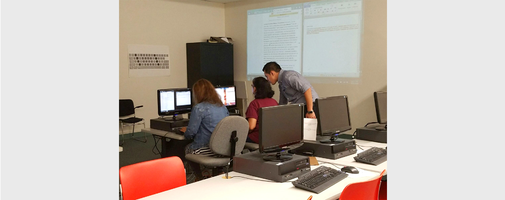 Photograph of three adults working at two stations in a computer lab.
