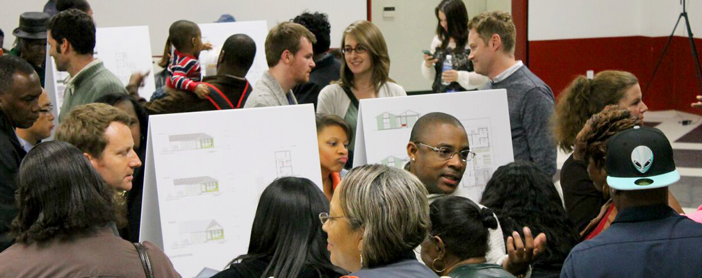 Photograph of two dozen residents in a large meeting room viewing and discussing posters of the housing designs set on easels.