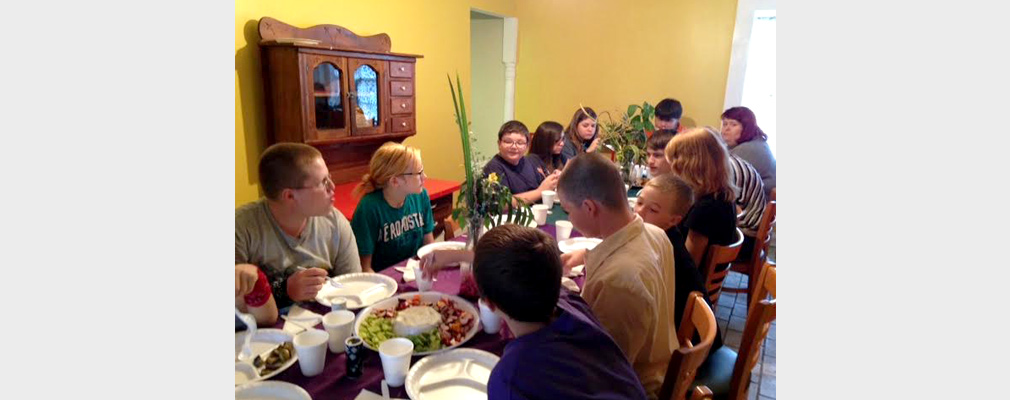 Interior photograph of a dozen children and adults eating lunch at a long table.