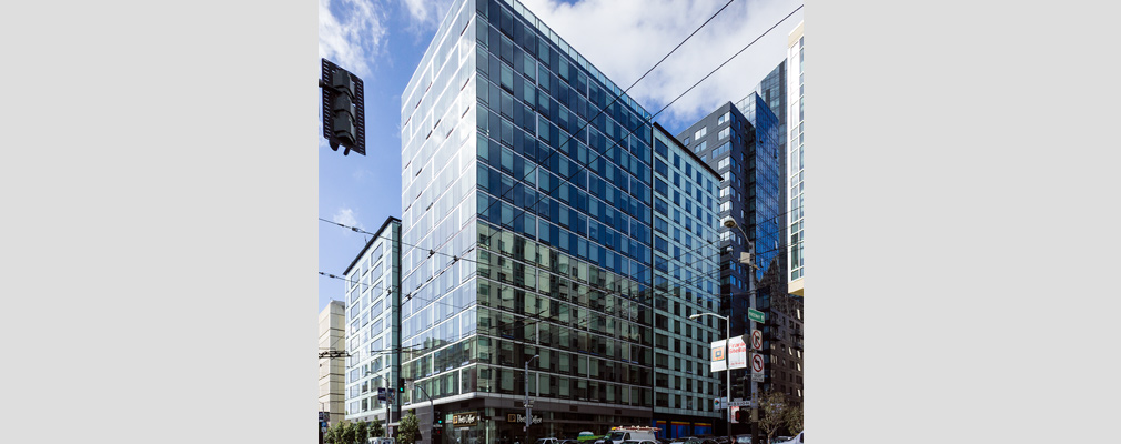 Photograph of the front and side façades of a 15-story building with commercial space on the ground floor and apartments on the upper floors; a 10-story section of the building is on the left side of the photograph.