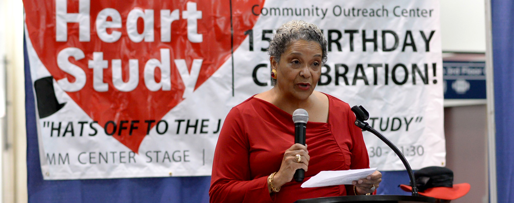 Photograph of a woman speaking with a microphone in front of a Jackson Heart Study banner.