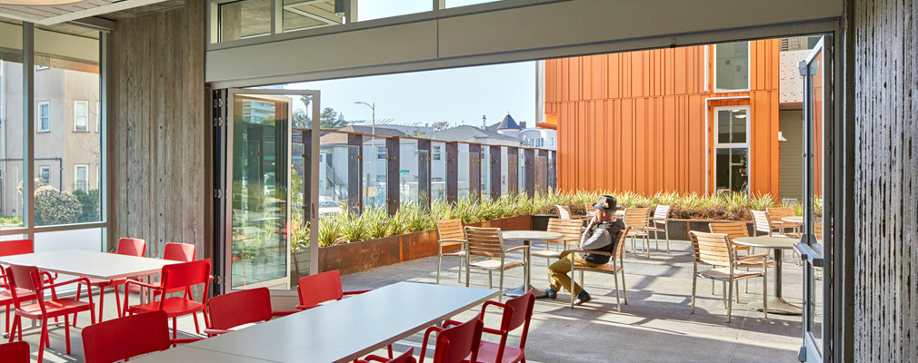 Photograph of an indoor lobby opened to a sunny outdoor courtyard separated from the street by a transparent glass wall.