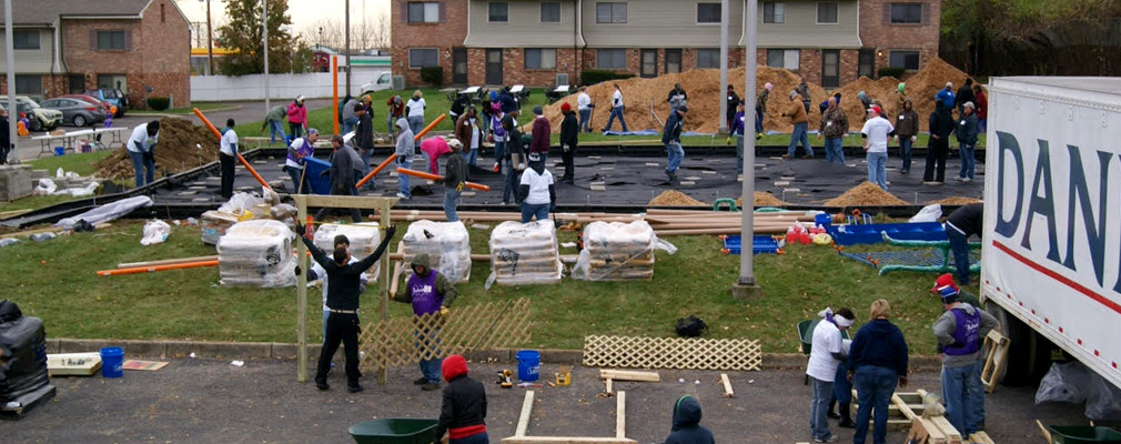 Photograph of a construction site with three dozen people, equipment, and materials in front of a two-story residential building. 