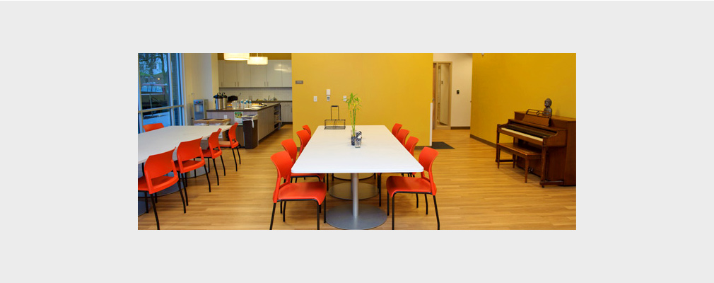 Photograph of a community room containing two long tables with chairs and an upright piano; a kitchen is in the background.