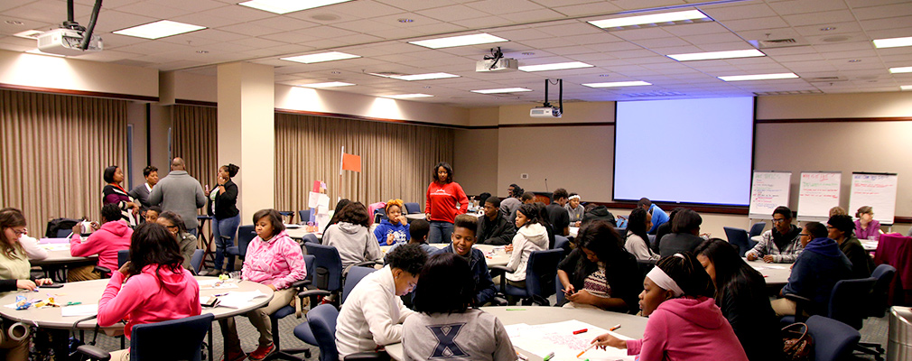 Photograph of a large meeting room with a projection screen and community members sitting at several round tables.