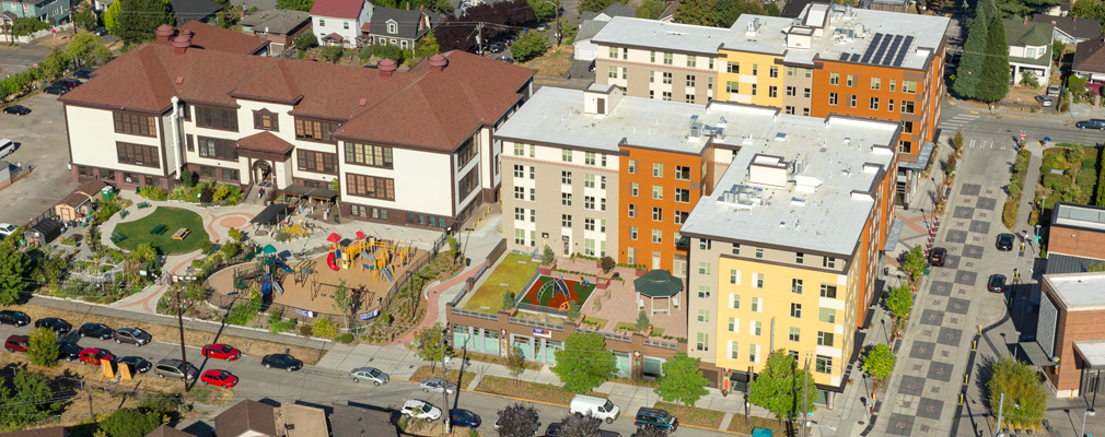 Low altitude aerial photograph of two 6-story buildings with a 3-story school to the left and a brick transit station on the right edge of the photograph.