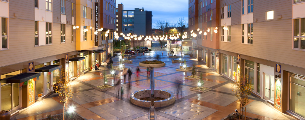 Photograph taken at dusk of a plaza framed on two sides by 6-story multi-use buildings.