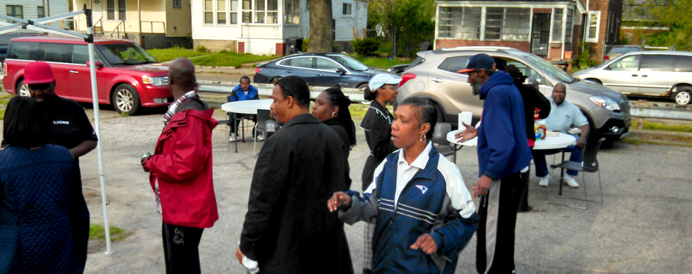 Photograph of a vacant lot where several men and women are standing and sitting beside tables.