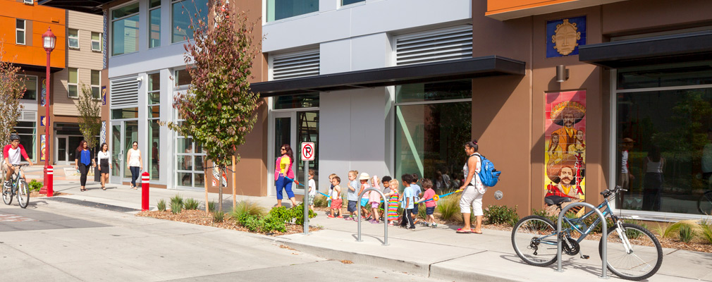 Photograph of a small group of young children accompanied by two adults in front of one of two multi-use buildings.