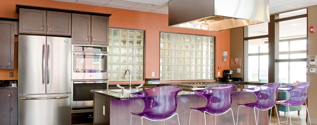 Photograph of demonstration kitchen with four chairs arranged along a kitchen island and with a stainless steel refrigerator and double oven in the background. 