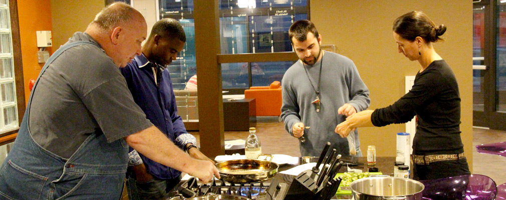 Photograph of three men and one woman cooking vegetables at a kitchen island. 