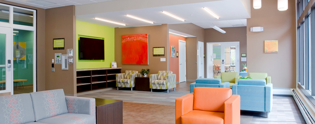 Photograph of a common area featuring couches and seven upholstered chairs; a glass door in the back wall separates this space from a meeting room. 