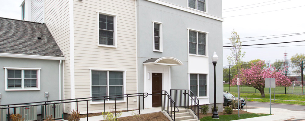 Photograph of the front façade of a three-story apartment building with a single-story wing on a street corner.