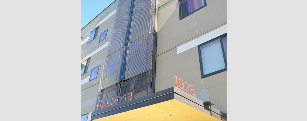 Photograph of an array of solar panels on the southern façade near the entrance to a four-story multifamily building.