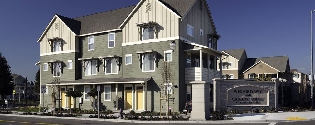 Photograph of the front and side façades of a 3-story multifamily building with wood siding and a pitched roof; a sign to the side of the building reads “Wexford Way and Carlow Court at Emerald Vista,” and similar buildings are in the background.