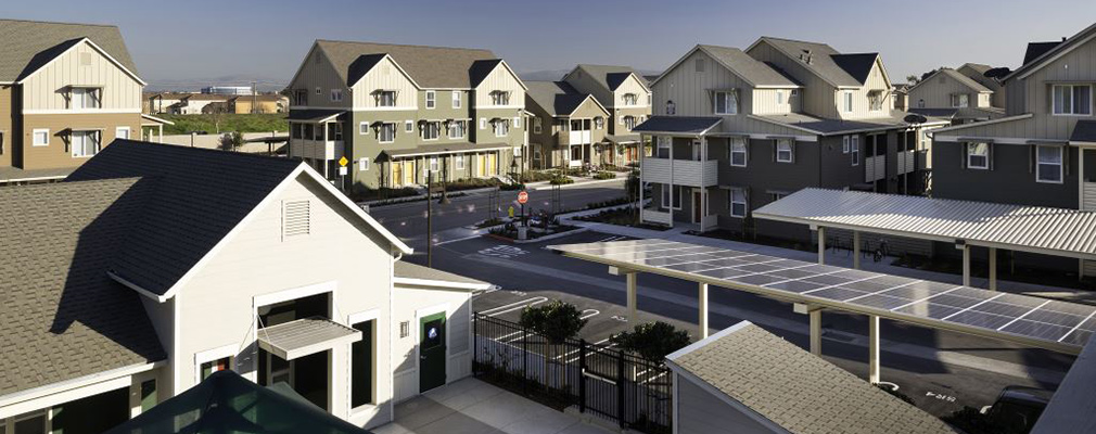 Photograph of parking spaces covered by a carport roof with solar panels; several multifamily buildings are in the background. 