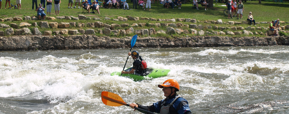 Photograph of two kayakers in whitewater with a large crowd watching from the riverbank in the background.