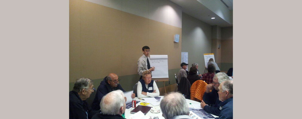 Photograph of residents at round tables in a large meeting room with students summarizing the discussions on easel pads.