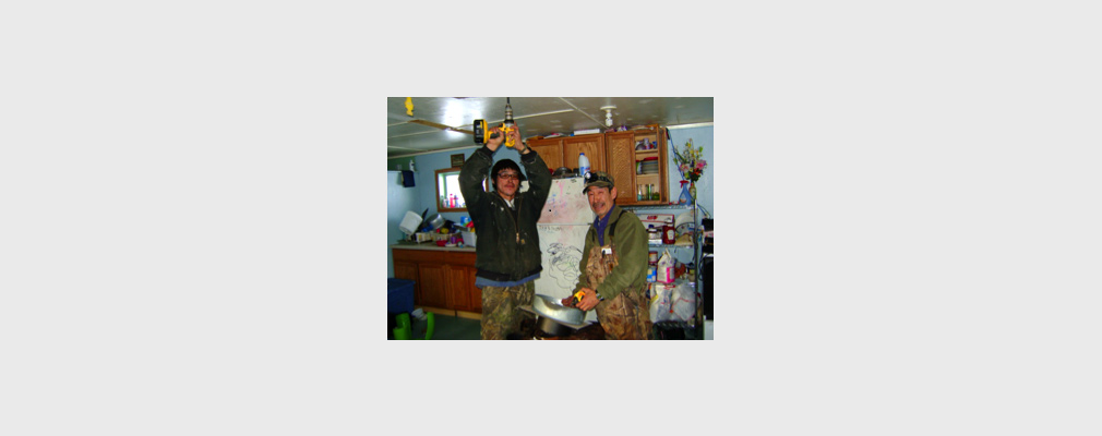 Photograph of two men in a kitchen drilling a hole in the ceiling for a chimney stack.