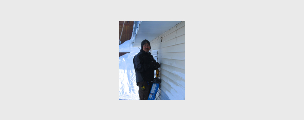 Photograph of a man standing on a ladder at the side of a house.