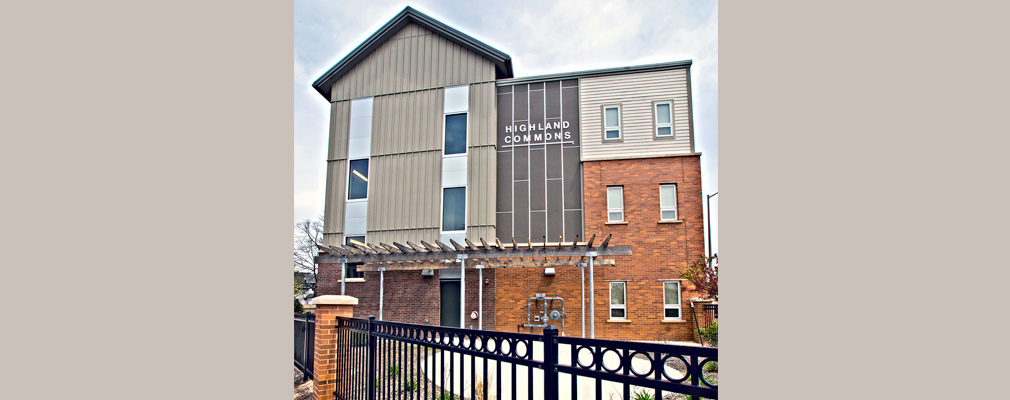 Photograph of a façade of a 3-story multifamily building standing behind a fenced yard with a patio.