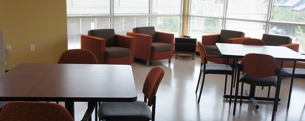 Photograph of a large room with two tables and sets of chairs and four upholstered chairs in front of windows along the back walls.