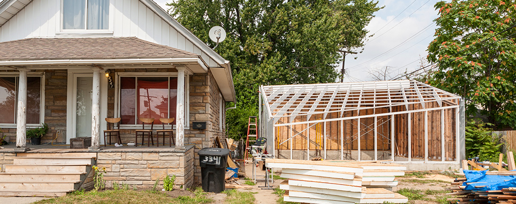 Photograph of a greenhouse under construction on a lot next to a house.