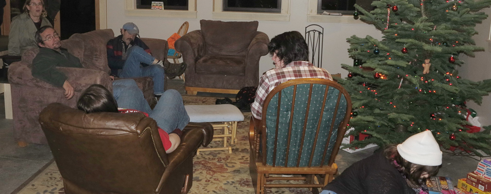 Photograph of six people in a large room with chairs, a couch, and presents around a Christmas tree.