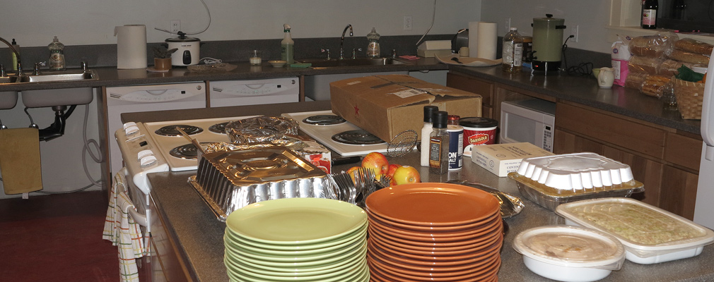 Photograph of a large kitchen with two stoves, two dishwashers, two sinks, and an island with several containers of prepared food and stacks of dishes.