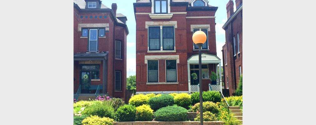 Photograph of a historic three-story, brick single-family house behind a small, landscaped front yard; portions of similar houses flanking the building are at the edges of the photograph.