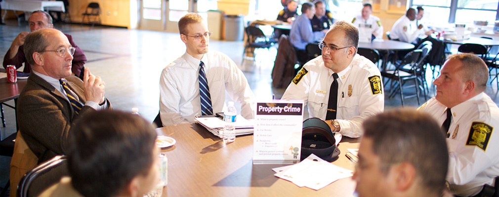 Photograph of police officers and other men sitting at several tables during a community meeting.