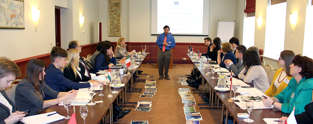 Photograph of a discussion leader standing in a large meeting room where approximately 20 people are seated at two long tables decorated with the flags of Lithuania, Poland, Spain, the United Kingdom, and other European countries.