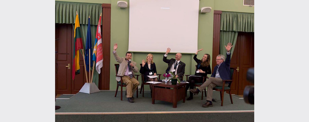 Photograph of five people seated at a small table on a stage participating in a show of hands with the audience during a presentation.