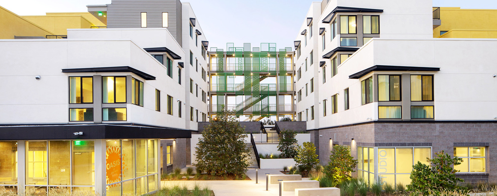 Photograph of two wings of a multistory residential building with groundfloor community rooms in the foreground and a raised courtyard accessible via stairs in the middleground.
